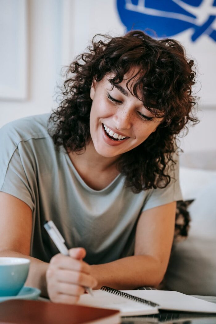 pexels-photo-6001386-6001386 Cheerful young female student with curly dark hair in casual clothes sitting near cute cat and taking notes while doing homework assignment